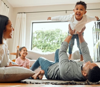 A happy family plays together indoors. A dad holds his son in the air while the mom and daughter watch with smiles.