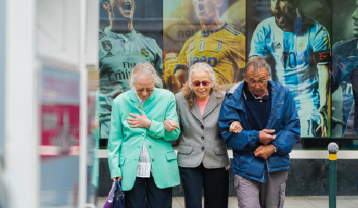Three elderly people walk arm in arm along a city street