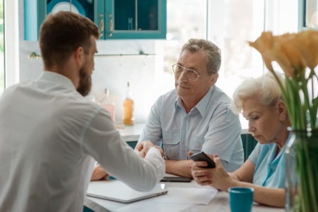 A young man in a white shirt shakes hands with a seated older man, as a seated older woman looks at her phone.